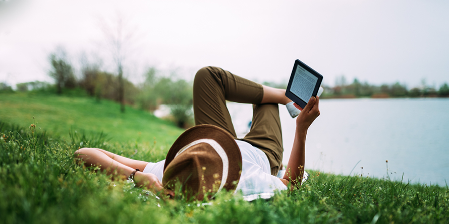 A person lays down on their back in the grass next to a lake with a brown hat on, reading an ebook. 